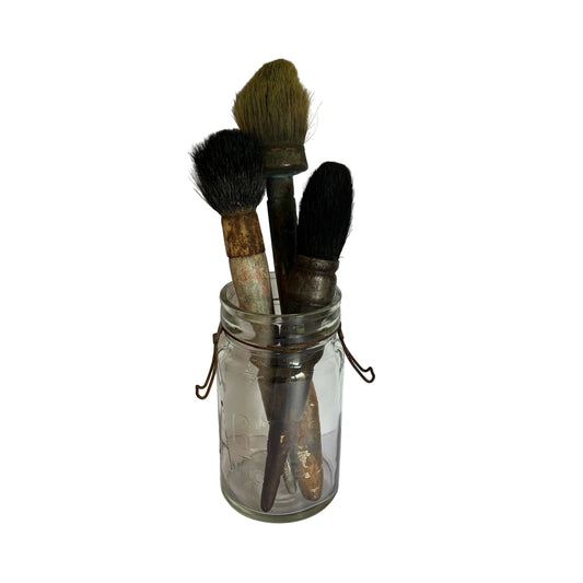 Three vintage makeup brushes with dark bristles and weathered handles, stored inside a clear glass jar with a metal clasp lid, set against a white paneled background.