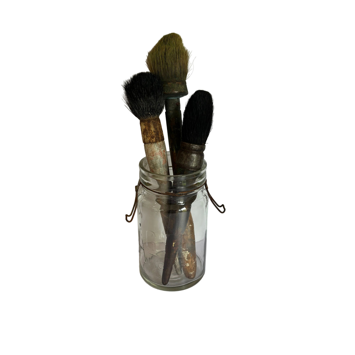 Three vintage makeup brushes with dark bristles and weathered handles, stored inside a clear glass jar with a metal clasp lid, set against a white paneled background.