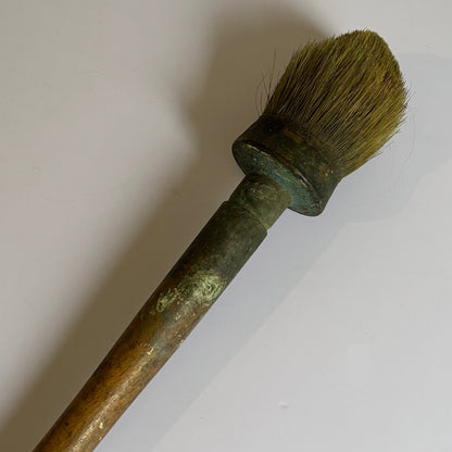 An antique-style paint brush with a long, weathered brown handle and a rounded, brush-like head in olive-green and brown tones, set against a plain off-white background.