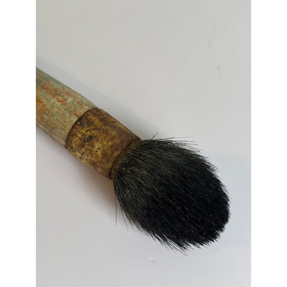 A close-up of a vintage makeup brush with a weathered, rusted metal ferrule and a light greenish-brown wooden handle, showing dense black bristles on a plain white background.